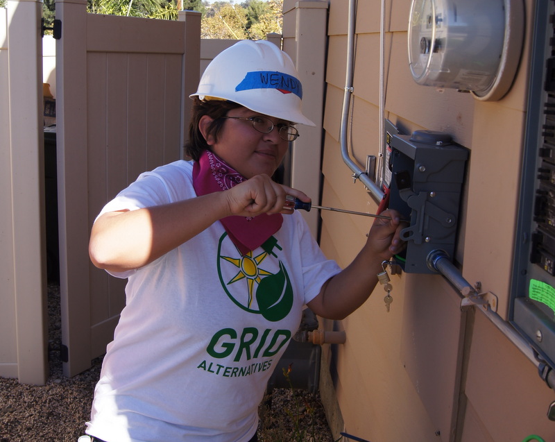 Worker in white hard hat and "Grid Alternatives" shirt installing solar equipment on residential meter box
