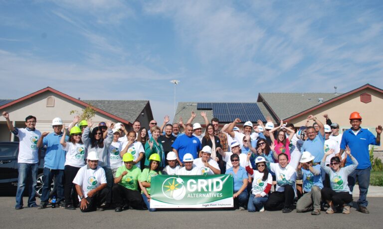 Large group of volunteers wearing hard hats and GRID Alternatives t-shirts holding green banner at solar installation site