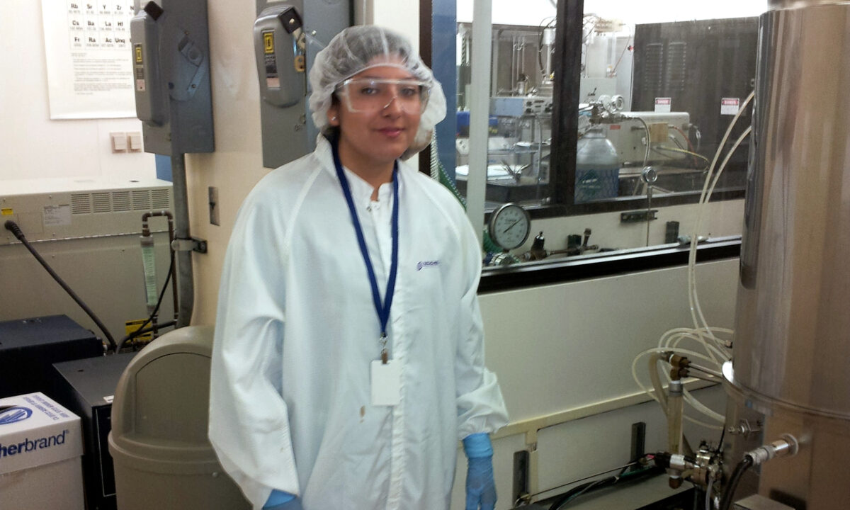 Woman in lab coat and safety gear stands in clean room laboratory with scientific equipment