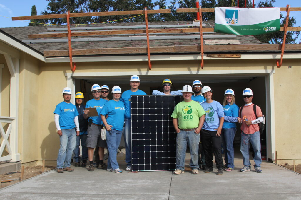 Team of volunteers in blue hard hats holding a large solar panel in front of a house under construction with solar equipment on the roof