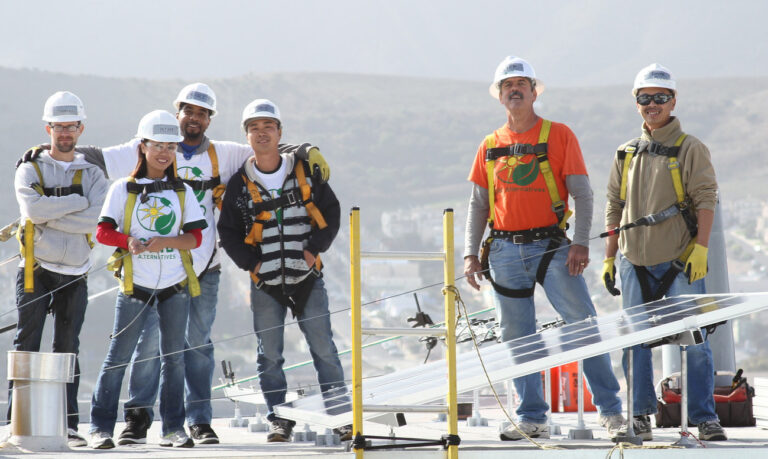 Six construction workers in hard hats and safety gear stand on a rooftop with city skyline behind them