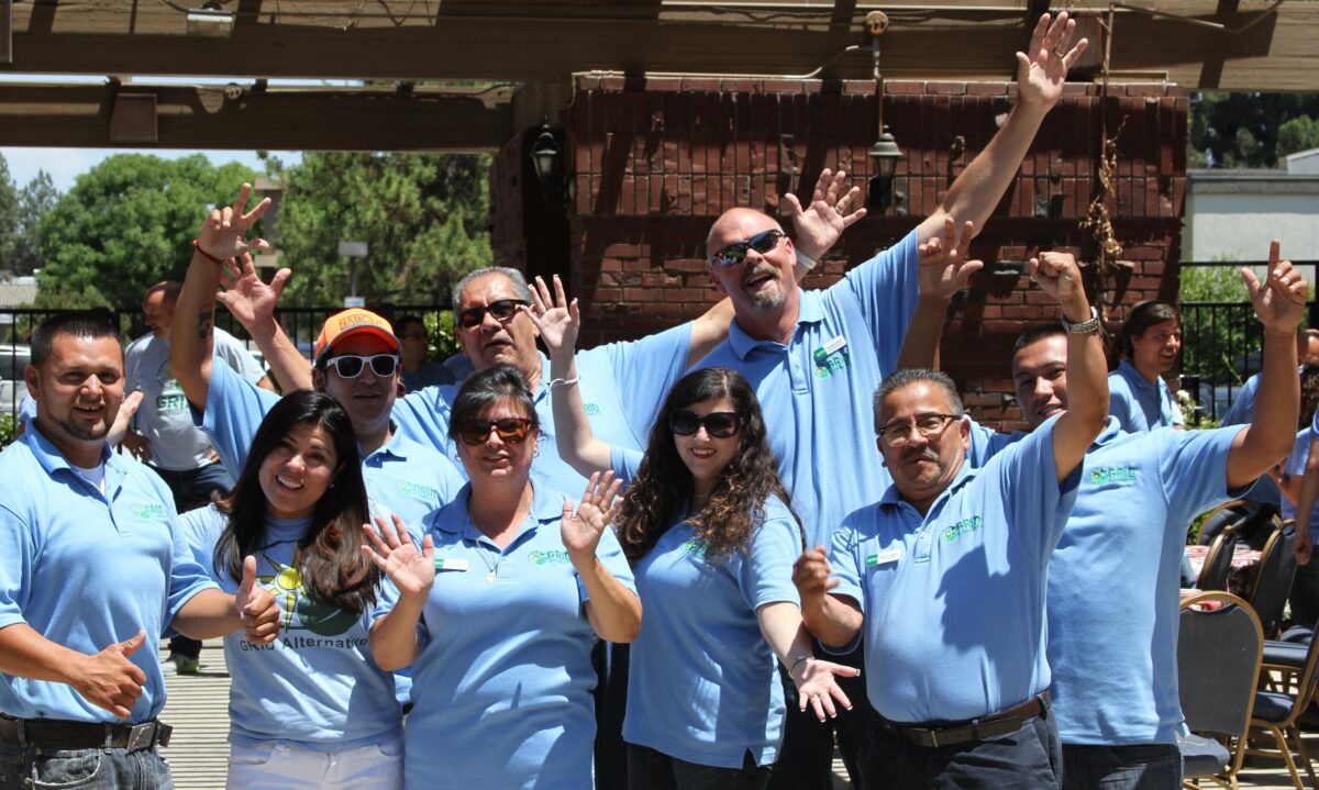 Group of team members in matching light blue shirts celebrating outdoors with raised arms