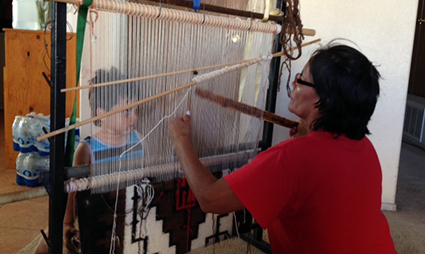Person in red shirt operating a large floor loom weaving at Ahidiikah Come Together event