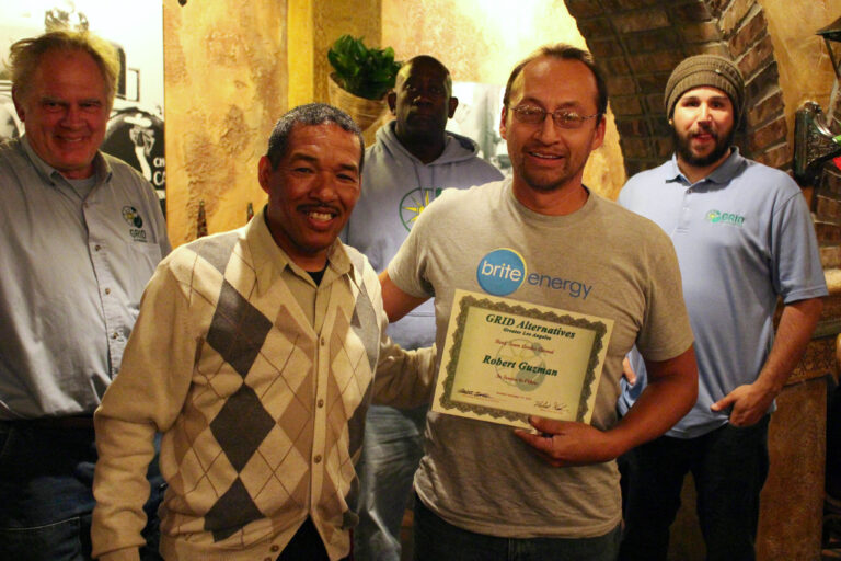 Five men posing together, center man holding a "Grid Alternatives" certificate for Robert Guchian, indoor setting with stone walls