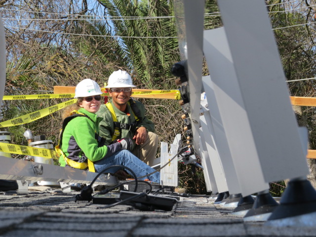 Two construction workers in hard hats and safety gear pose on a rooftop with yellow caution tape visible behind them
