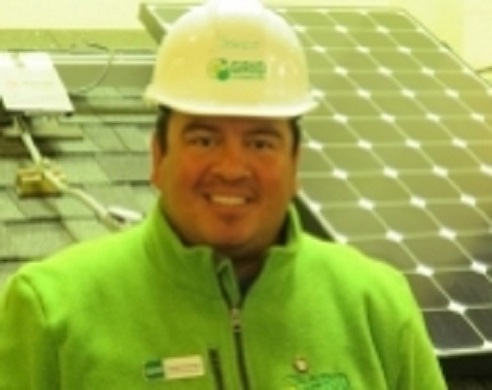 Sergio Coronel smiling in safety gear with white hard hat and neon green jacket near solar panels