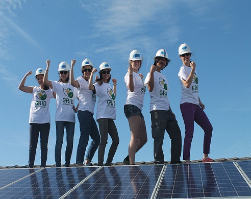 Group of nine people in white hard hats and "GRID" t-shirts standing on solar panels with arms raised