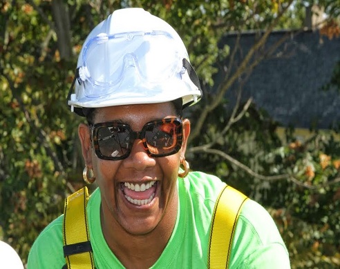 Smiling woman in white hard hat, green shirt, and safety vest outdoors