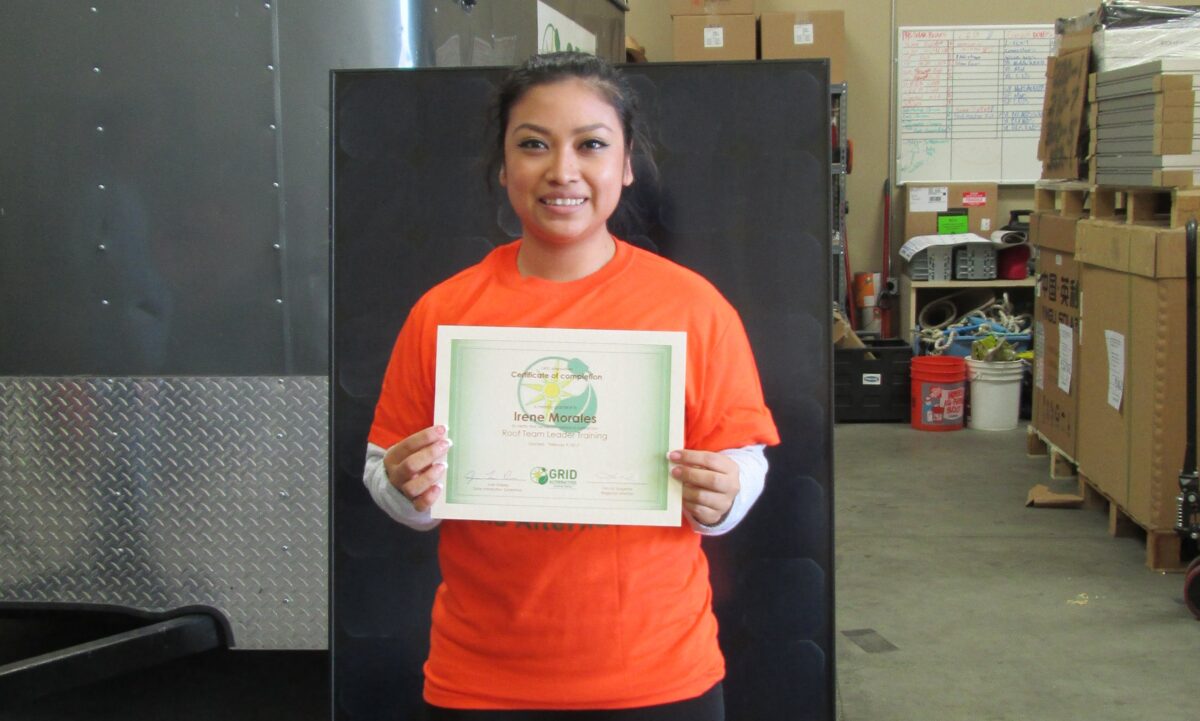 Woman in orange shirt holding certificate of recognition for Irene Morales in warehouse setting