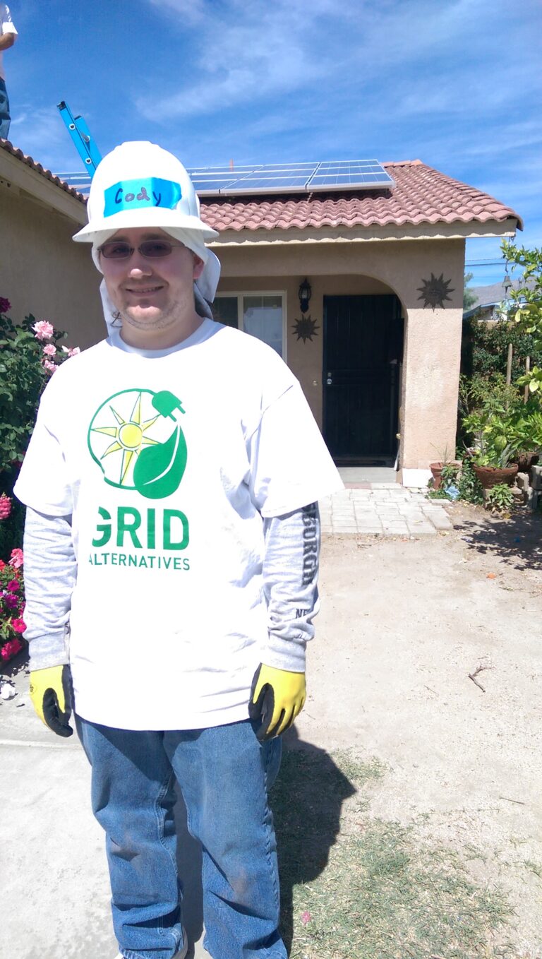 Man wearing GRID Alternatives shirt and hard hat standing in front of residential home with solar panels