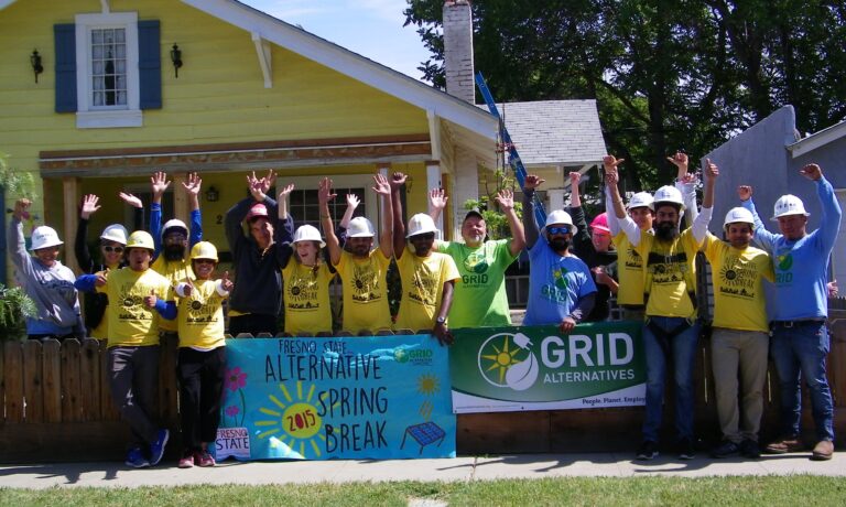Volunteers in yellow shirts and hard hats celebrate outside a yellow house during Fresno State Alternative Spring Break 2015 with GRID Alternatives banner