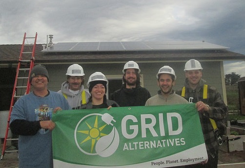 Six workers in hard hats holding a GRID Alternatives banner in front of a house with solar panels on the roof