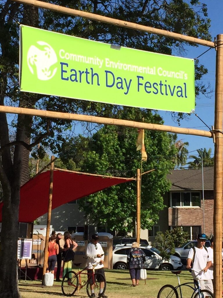 Bright green banner reading "Community Environmental Council's Earth Day Festival" at outdoor event with attendees and bicycles