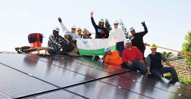 Group of workers in hard hats celebrating on rooftop with solar panels and company banner