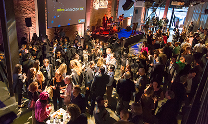 Crowded indoor event with attendees mingling, stage with projection screen visible in background