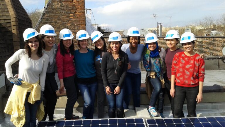 Group of women wearing white hard hats and blue logos on a rooftop with solar panel, smiling at camera