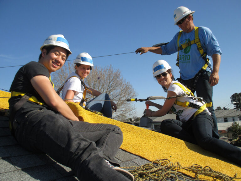 Five people in safety gear and harnesses install yellow solar panels on a roof under clear blue sky