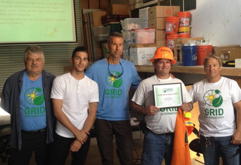 Five men wearing GRID Alternatives shirts, one holding a Certificate of Appreciation, standing in a workshop with tools and supplies