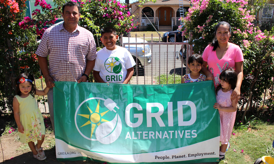 Family holding GRID Alternatives banner with sun logo in front of blooming magenta flowers and house