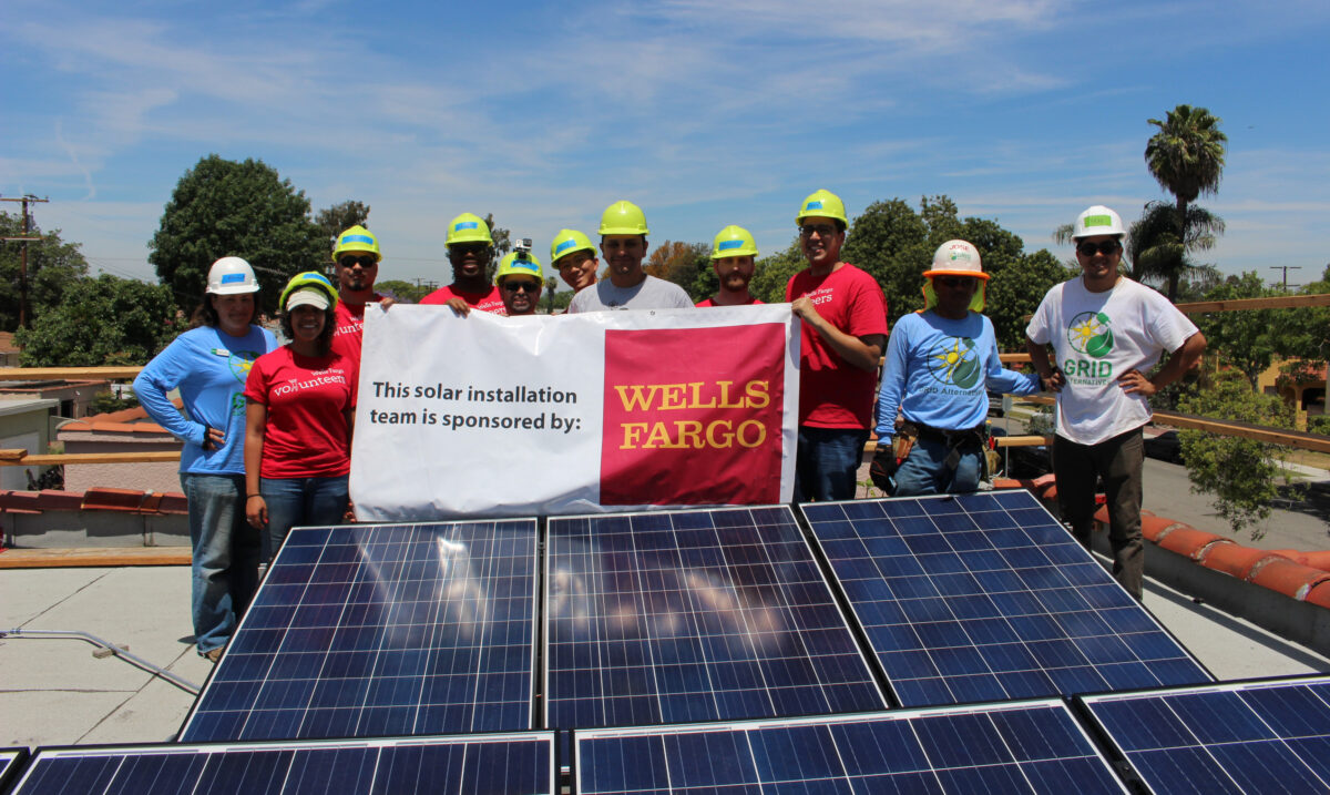 Solar installation team sponsored by Wells Fargo posing on rooftop with solar panels and banners