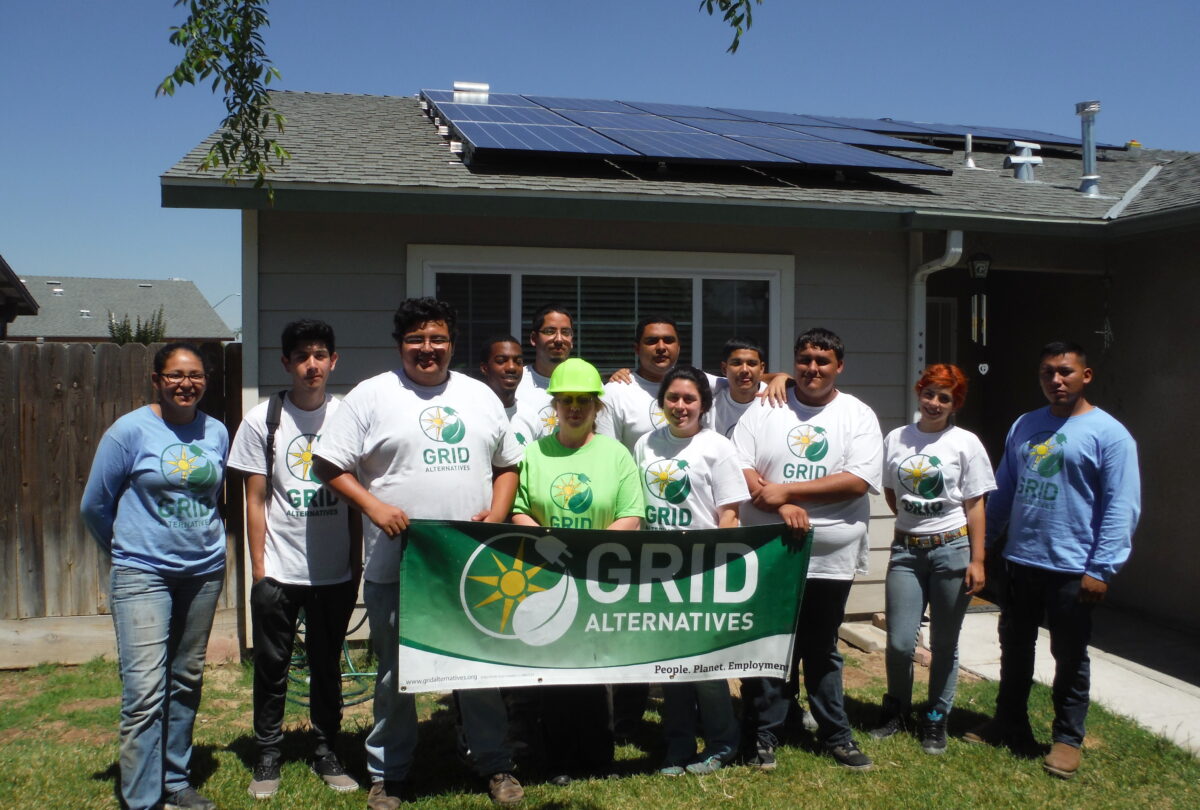 Group of GRID Alternatives staff and volunteers holding banner in front of house with solar panels