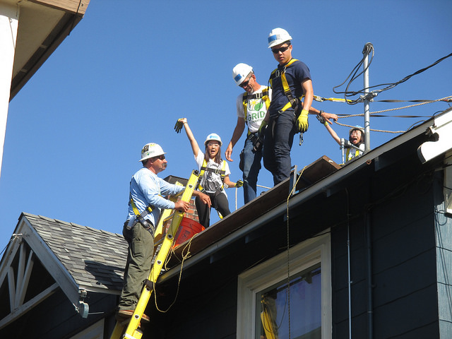 Construction workers on ladder and roof installing solar panels on residential house under clear blue sky