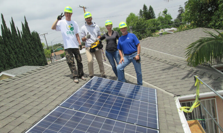 Four workers in safety helmets stand on a residential roof next to a newly installed solar panel