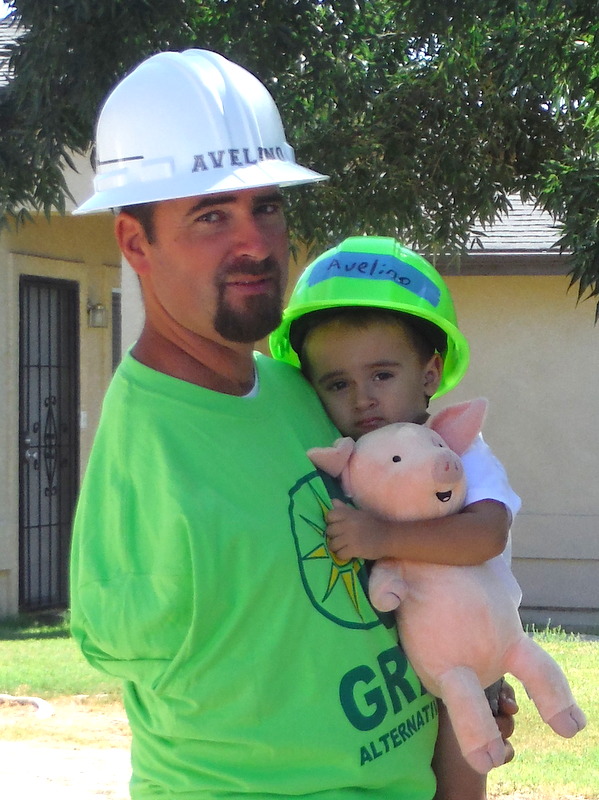 Man and child wearing hard hats labeled "Avelino" pose outdoors, child holding stuffed pig toy