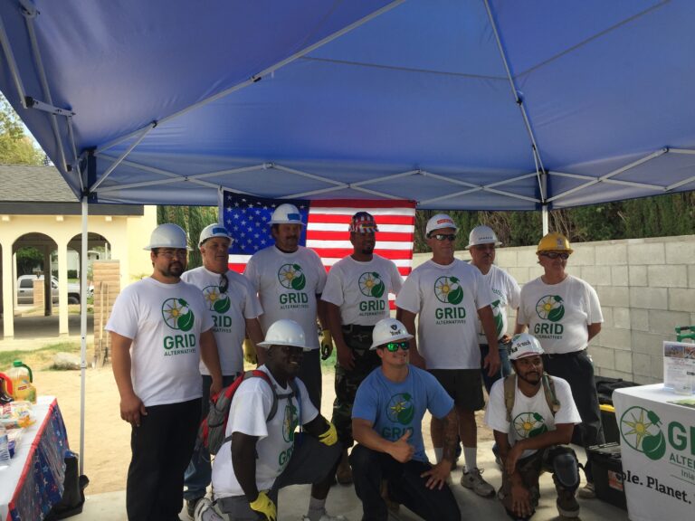 Group of workers in GRID Alternatives shirts and hard hats under blue tent with American flag