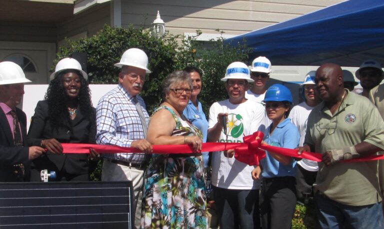 Group at ribbon-cutting ceremony wearing hard hats, holding red ribbon with official attendees