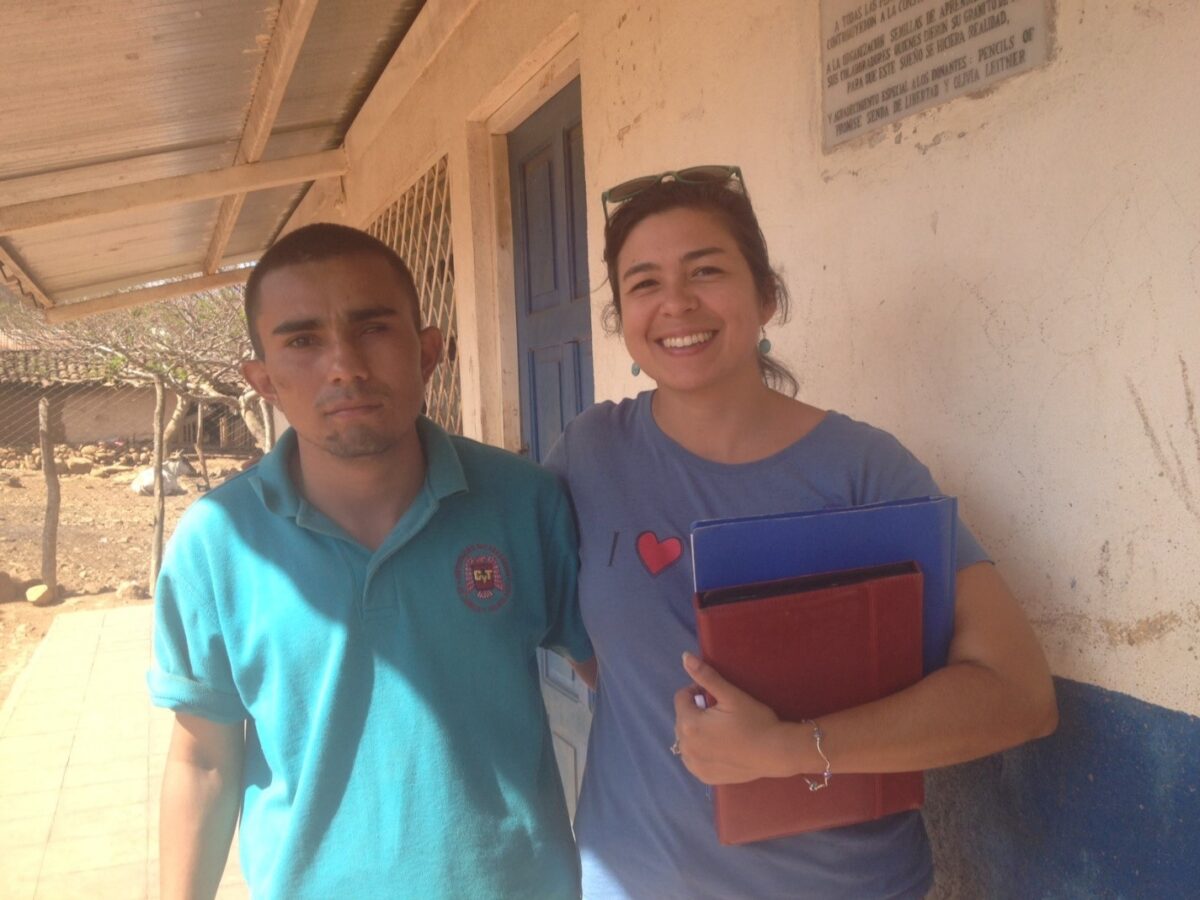 Two people standing together outside a building, one wearing a turquoise polo shirt and one holding a blue and red folder, both smiling at the camera
