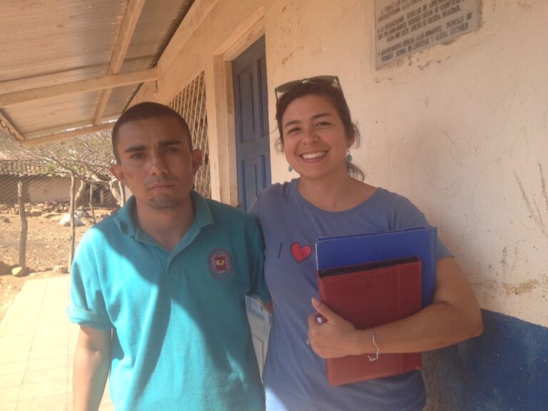 Two people standing together outside a building, one wearing a turquoise polo shirt and one holding a blue and red folder, both smiling at the camera