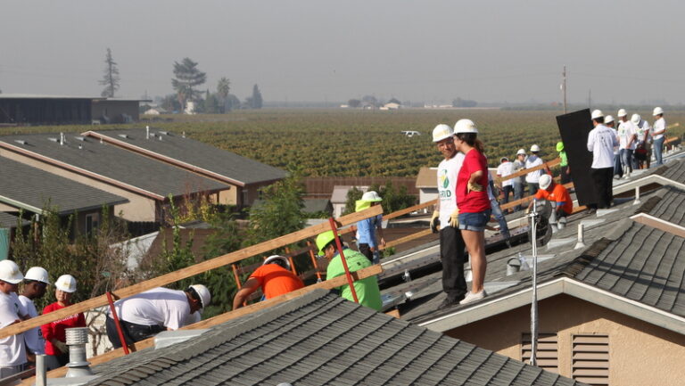 Construction workers in hard hats on residential roofing project with agricultural fields visible in background