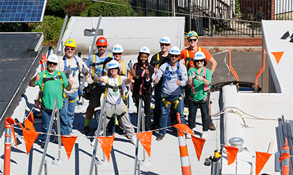 Group of construction workers and professionals wearing hard hats at a building dedication event with orange and white bunting