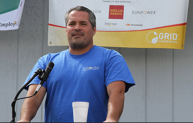 Man in blue shirt speaking at microphone in front of sponsor banner