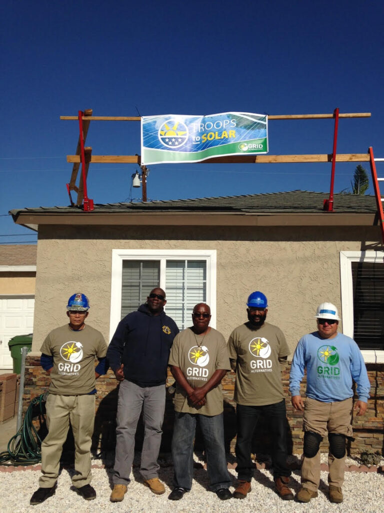 Five workers in hard hats and GRID shirts stand in front of a house with "Troops to Solar" banner on roof structure