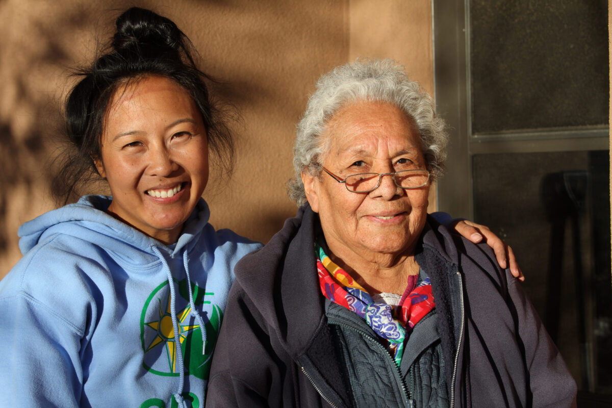 Two women smiling together outdoors, one younger in a blue hoodie and one elderly in a dark jacket with colorful scarf