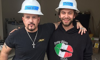 Two men wearing white hard hats and black shirts pose together indoors, smiling at the camera