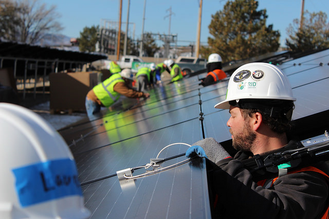 Worker installing solar panels on rooftop with construction crew in background