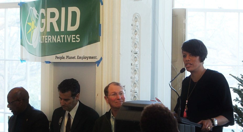 Woman speaking at podium under GRID Alternatives banner with four men seated behind her