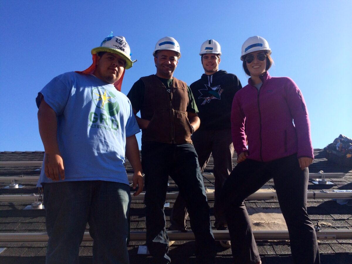 Four construction workers wearing hard hats stand together on a roofing site under clear blue sky
