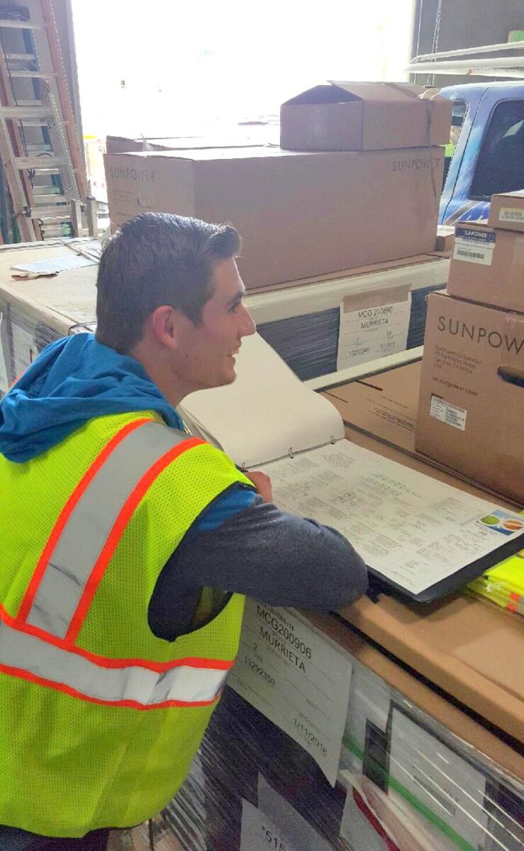 Worker in safety vest reviewing shipping documents next to cardboard boxes labeled "Sunpower" on a truck bed