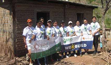 Group of volunteers in white shirts holding "We Build" banner in front of wooden structure
