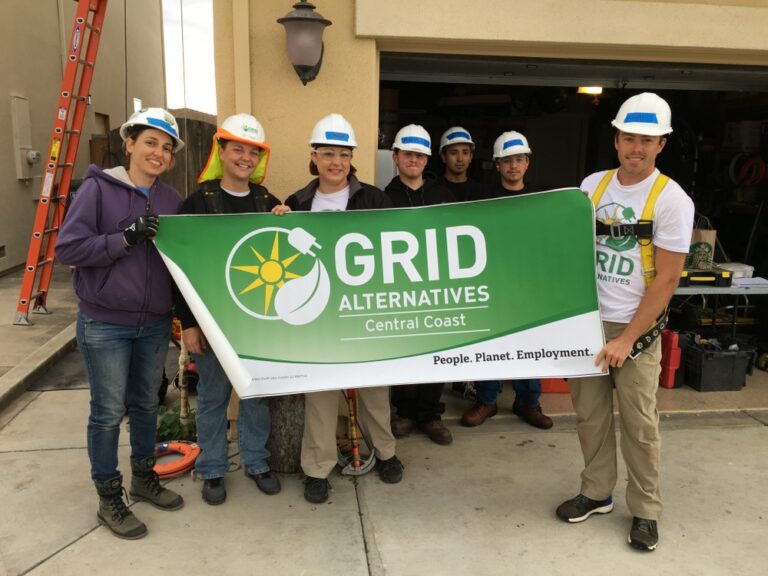 Seven people in hard hats holding a green GRID Alternatives Central Coast banner on a construction site