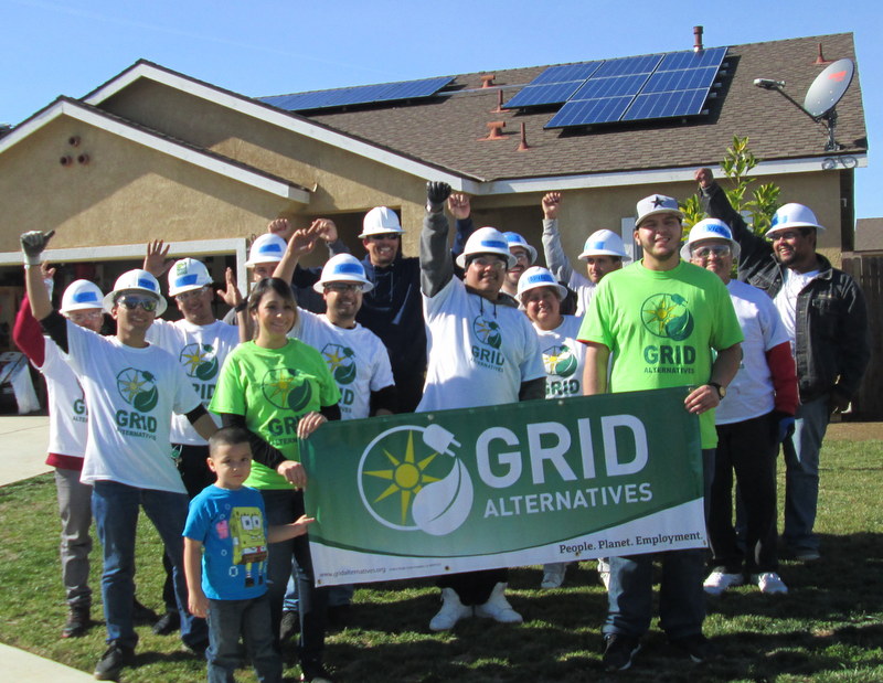 Group of volunteers in hard hats holding GRID Alternatives banner in front of house with solar panels