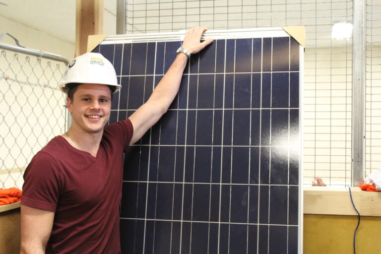 Young man in hard hat and maroon shirt holds a solar panel and smiles at camera