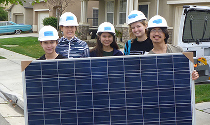 Five people wearing hard hats hold a large solar panel in front of a house