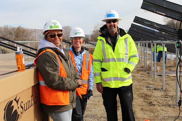 Three construction workers in safety gear and hard hats stand together on a solar panel installation site