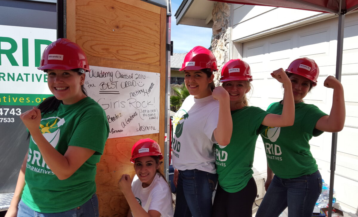 Six students in red hard hats and green "GRID Alternatives" shirts pose flexibly at a construction site, smiling at the camera