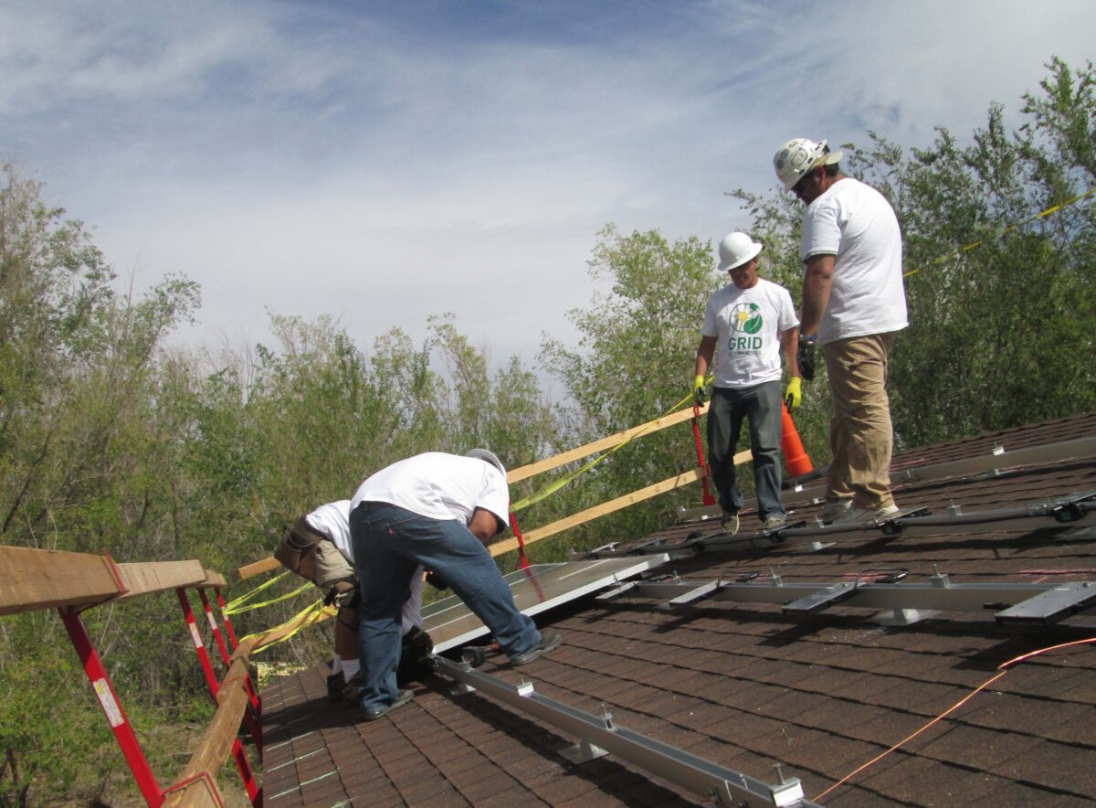 Three workers in hard hats and safety gear installing solar panels on a residential roof with green trees in background